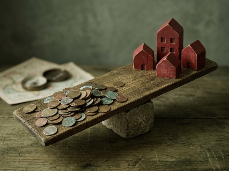 Wooden seesaw balancing a pile of coins against small red model houses