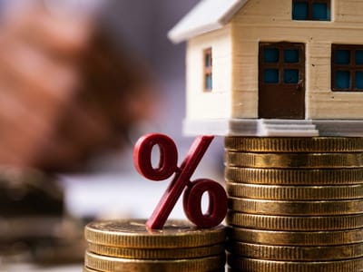 Red percent sign leaning on stacks of coins beside a model house