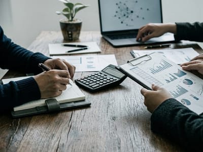 Two people reviewing bar charts with a calculator and notebook at a wooden desk
