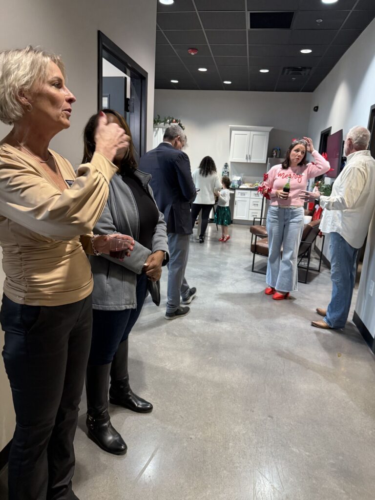 Guests mingling at an office Christmas party, with a woman in a pink Merry sweater holding a bottle and others conversing near a kitchenette decorated for the holidays