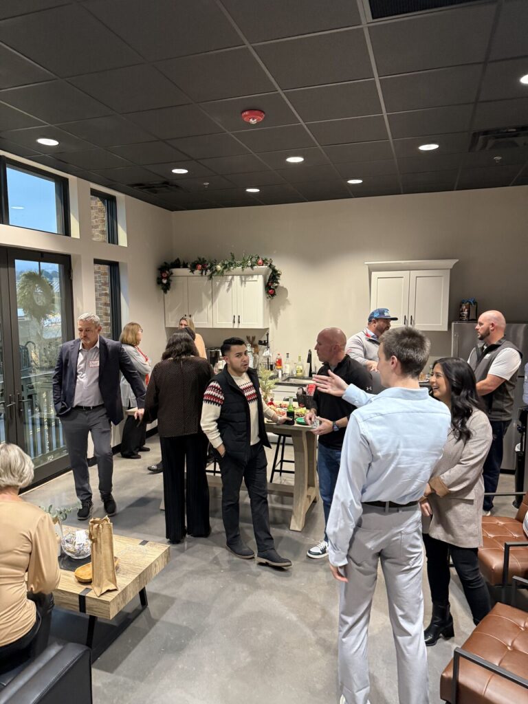 Colleagues mingling at an indoor office Christmas party, with holiday garland above the kitchen cabinets and gift bags on a low table in the foreground