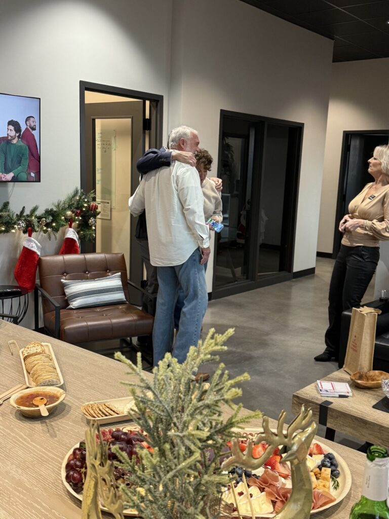 Two guests hugging at an office Christmas party, with a charcuterie board and gold reindeer centerpiece in the foreground and holiday garland with stockings in the background