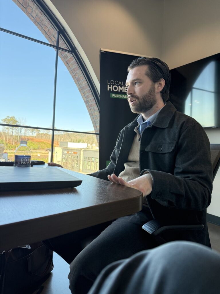 Adam Buice seated at a table during a meeting, speaking with hands clasped, Local Home Purchasers banner and large arched window visible behind him